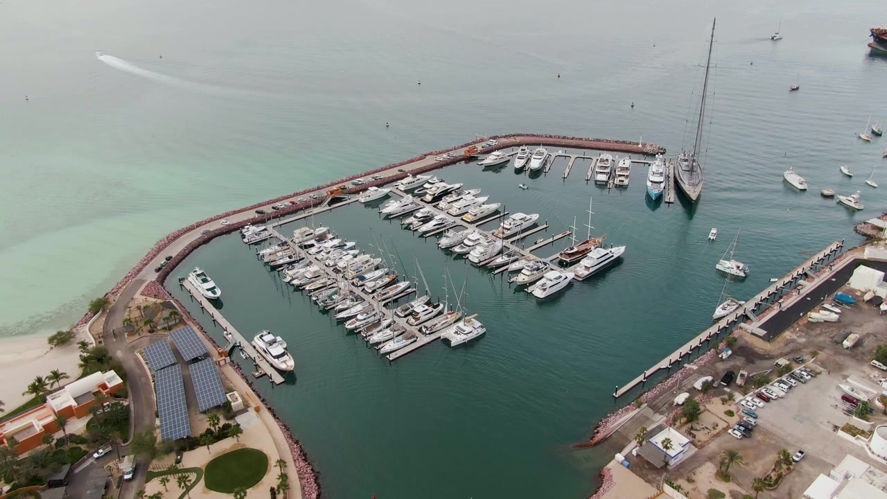 Aerial view of the La Paz marina one second in, with the docked boats filling the basin, larger sailboats near the right breakwater, shoreline buildings below, and no visible people.
