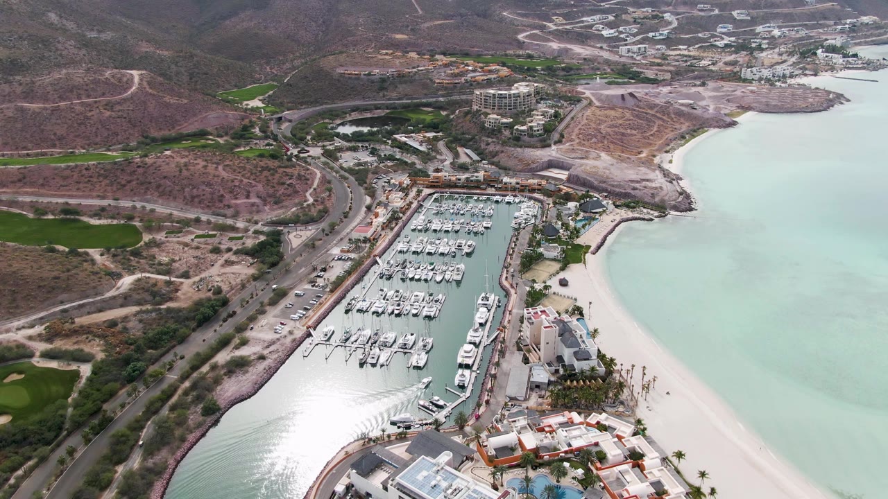 At 0:02, the La Paz aerial view shows the marina slightly farther across frame, with clustered white boats, shoreline sand, pale water, nearby buildings, roads, arid hills, and no visible people.