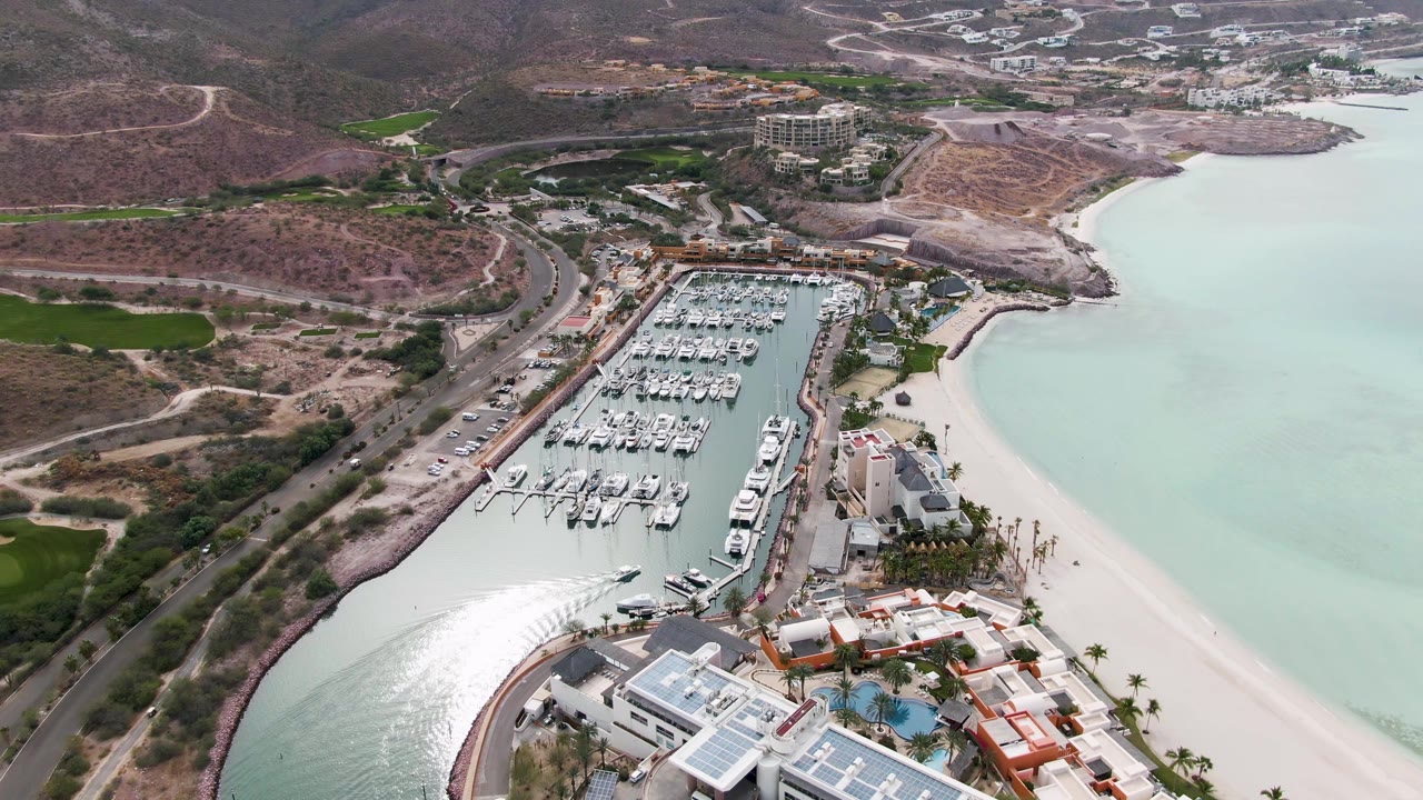 Aerial frame over La Paz, Baja California Sur showing docked boats in a marina beside a sandy beach, pale green water, hillside roads, and no visible people.