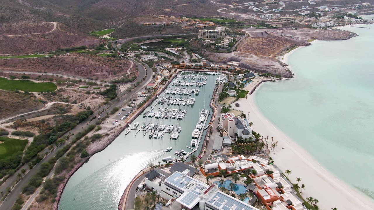 La Paz aerial frame showing rows of white boats in the marina, resort-like buildings near the beach, pale coastal water, hills beyond, and no visible people.