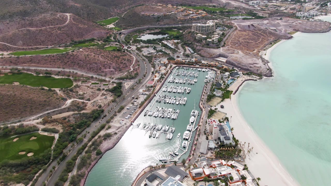 At 0:02, the drone frame keeps the marina centered while a coastal road curves past dry hills and golf fairways, with pale beach and turquoise water to the right and no visible people.