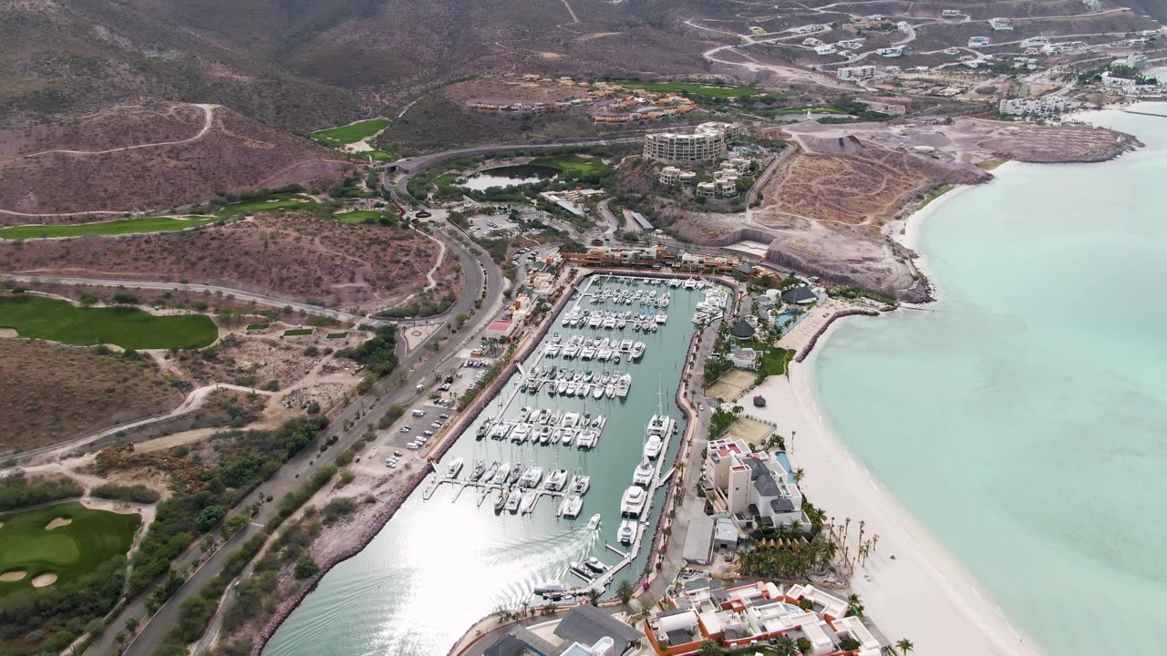 High aerial frame from La Paz, Baja California Sur with docked boats centered in the marina, bright glare near the channel, a sandy beach to the right, golf greens to the left, and no visible people.