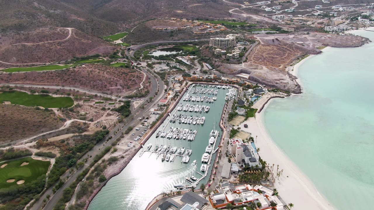 Drone-style aerial frame over La Paz, Baja California Sur with the marina shifted lower in view, pale water along the beach, green golf areas at left, hillside roads and buildings beyond, and no visible people.