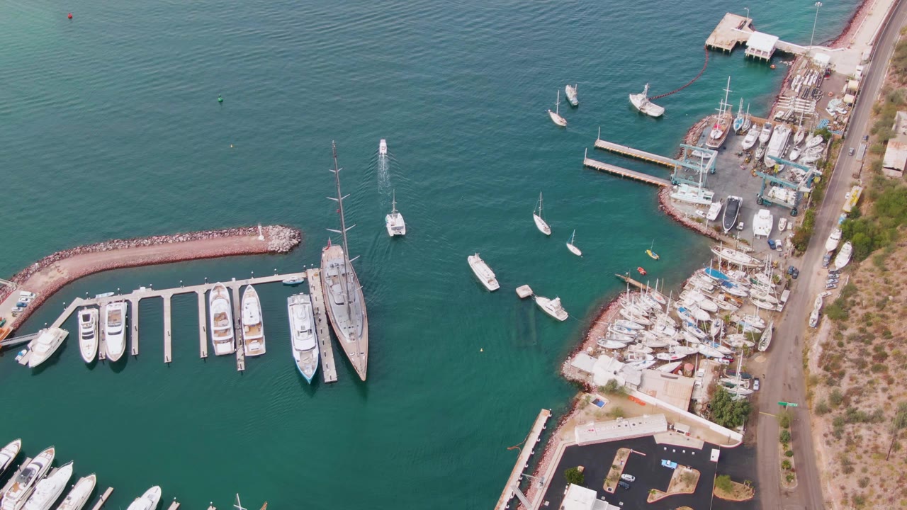 At 0:02, the aerial view shows the curved breakwater, rows of docked boats, a large sailboat at the marina edge, shoreline work areas, and no visible people in La Paz.