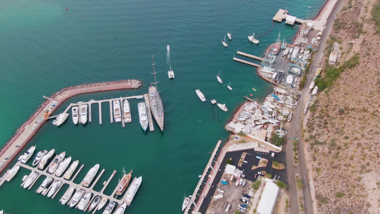 Aerial frame over La Paz marina showing white yachts in slips, small vessels in open turquoise water, a shoreline work area, a road, dry coastal land, and no visible people.