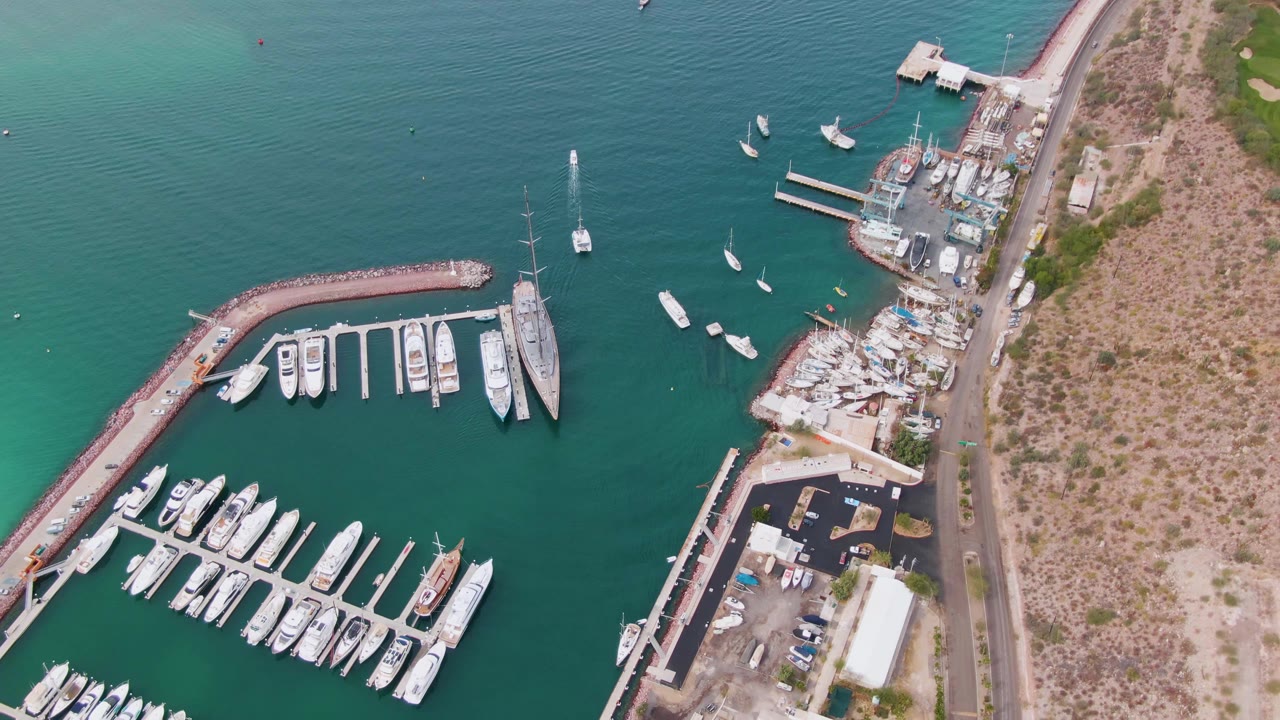 High drone view in La Paz, Baja California Sur with docked boats arranged in marina slips, a red-toned curved breakwater, moored sailboats, a road along dry terrain, and no visible people.