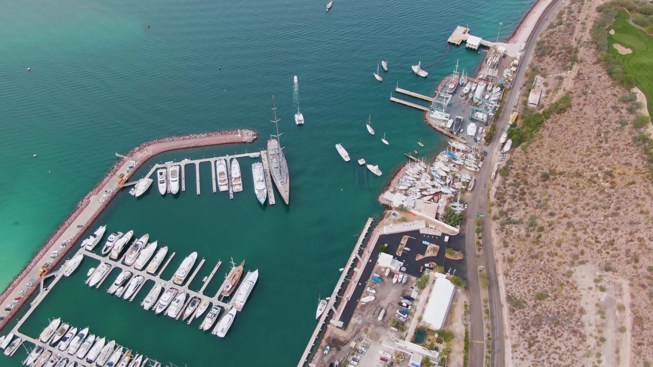 Top-down La Paz marina view with rows of docked boats, piers, turquoise water, a road beside dry hillside, a green landscaped area at the upper right, and no visible people.