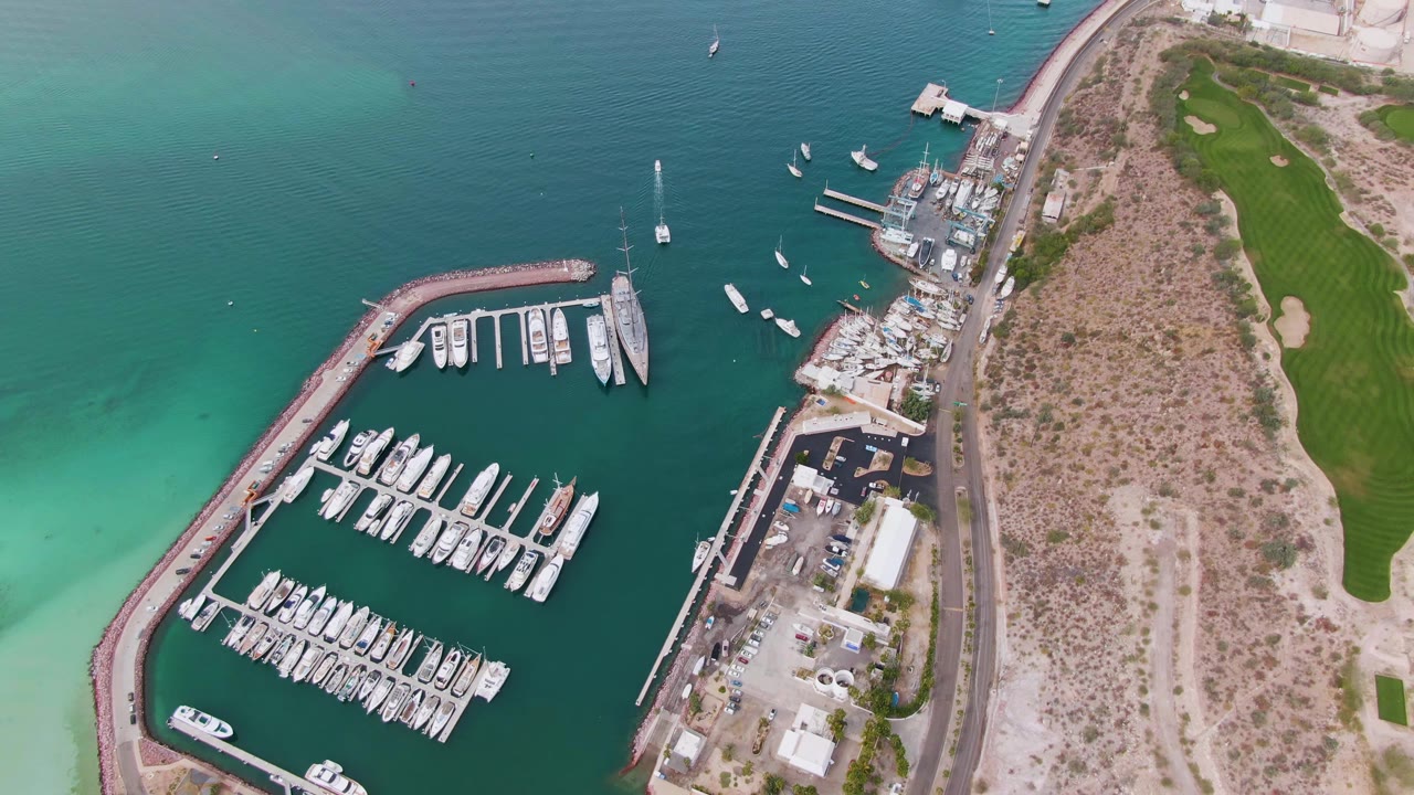 Opening overhead frame of a La Paz, Baja California Sur marina with docked white boats, a curved breakwater, a shoreline road, green grass, and no visible people.