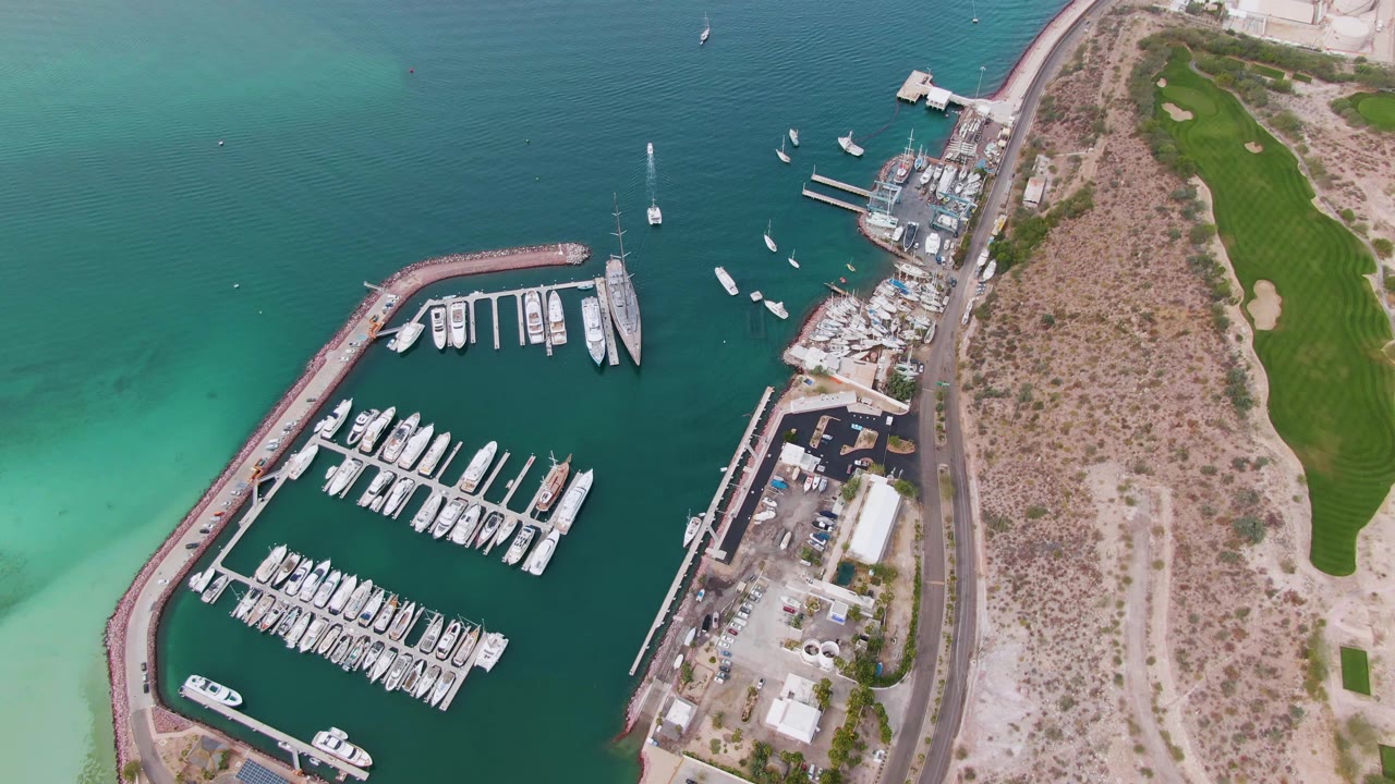 One second into the clip, the La Paz marina remains centered with rows of white boats, deeper blue water outside the docks, a road along the shore, and no visible people.