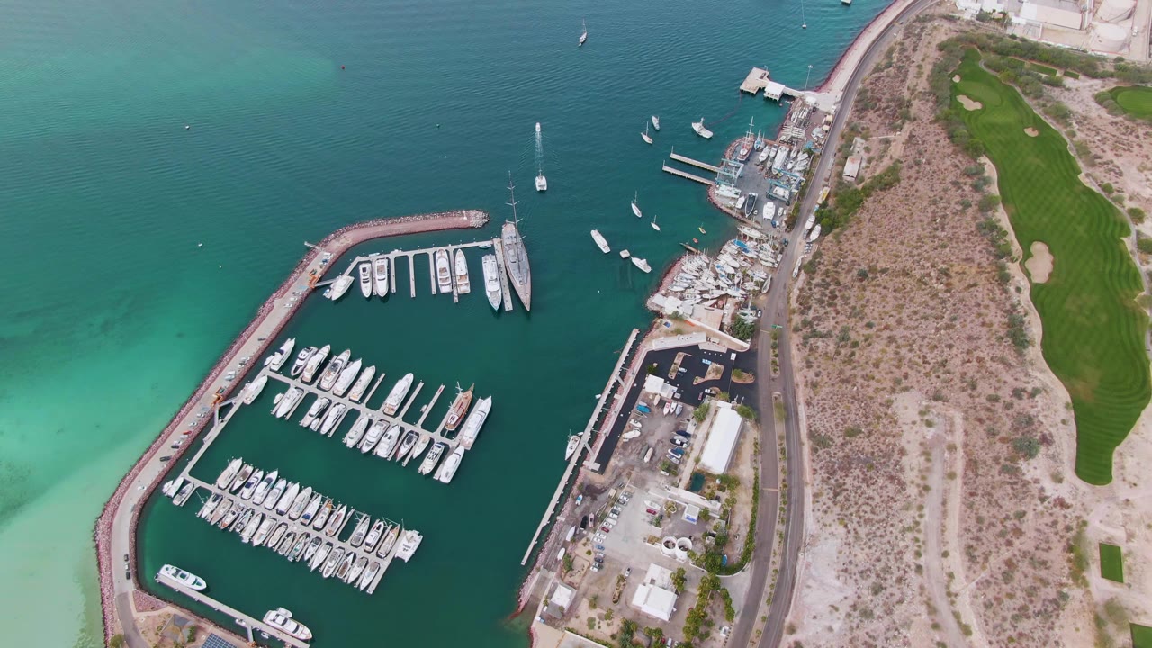 At 0:03, the La Paz marina is seen from above with clustered docked boats, pale turquoise water near the breakwater, shoreline buildings, a road, and no visible people.