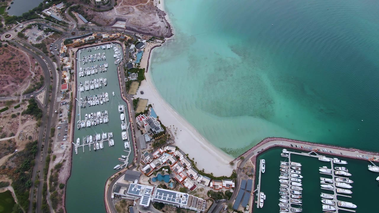 Aerial frame over La Paz, Baja California Sur showing two marina basins with rows of boats, a curved sandy beach, turquoise shallows, nearby roads, and no visible people.
