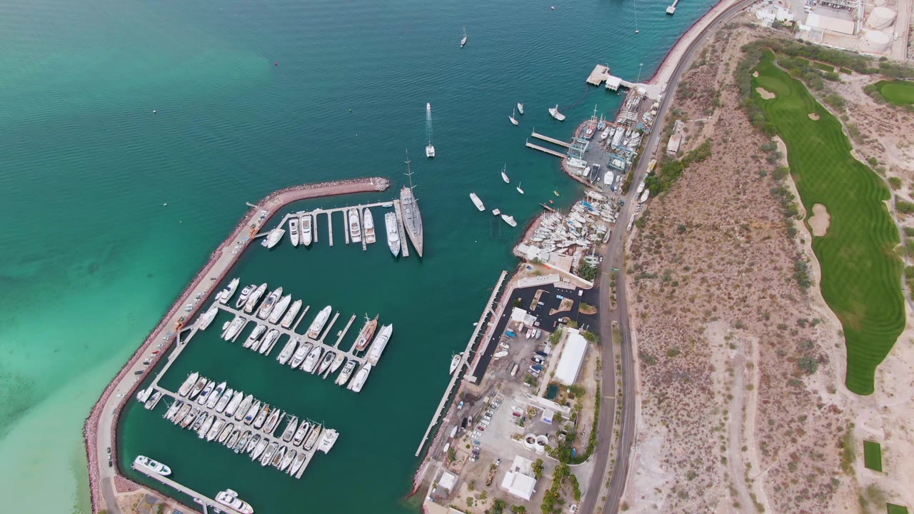 Overhead marina with docked boats, turquoise water, coastal road, and golf course edge.