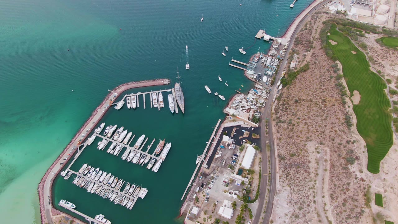 Aerial frame from La Paz with docked boats in the marina, small boats on the surrounding water, the coastal road through dry waterfront land, golf course grass to the right, and no visible people.