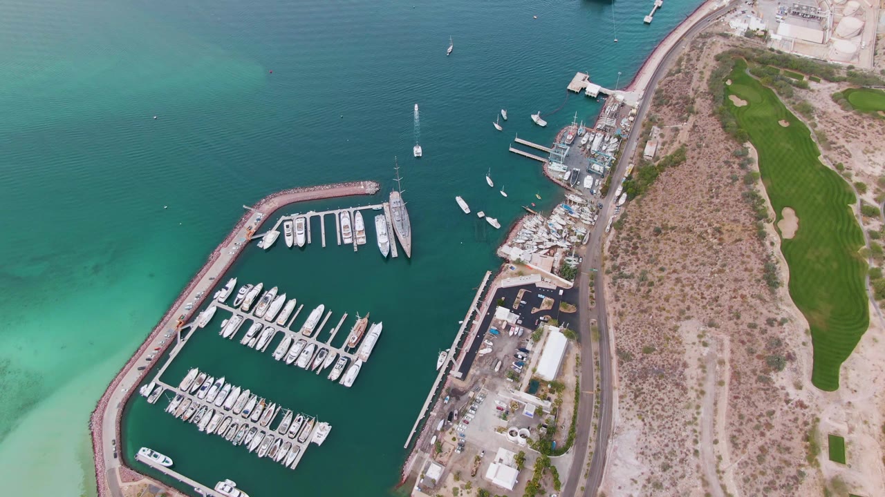 Overhead frame in La Paz showing bright shallow turquoise water at left, dark marina water with docked boats below, a waterfront road and dry land at center, golf course grass at right, and no visible people.