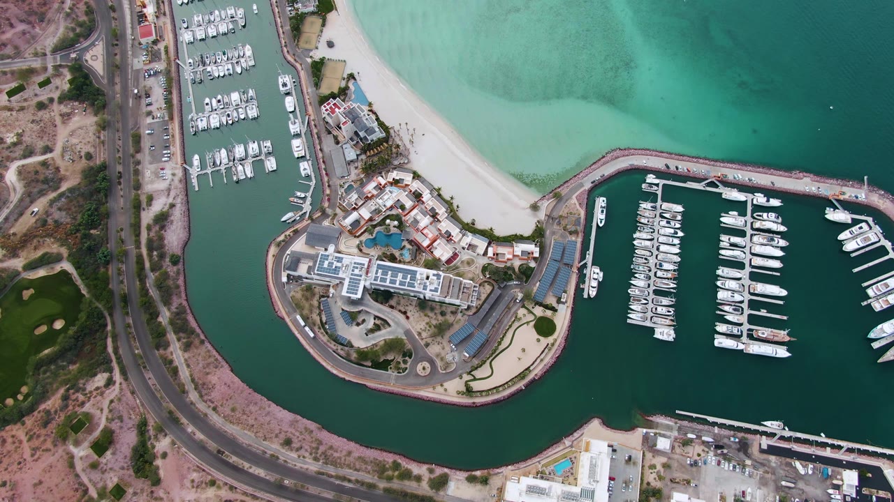 High aerial frame over La Paz showing marina slips to the right, pale sand and shallow turquoise water above, roadside dry terrain to the left, and no visible people.