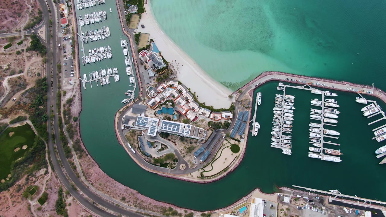 Aerial marina scene with road, dry hillside, buildings, beach, and docked boats.