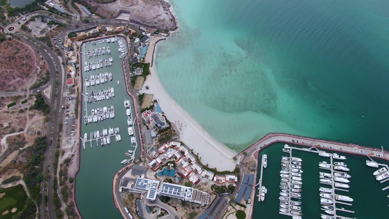 High drone view over La Paz with two marina basins, clustered white boats, a waterfront building area, pale beach sand, green-blue water gradients, and no visible people.