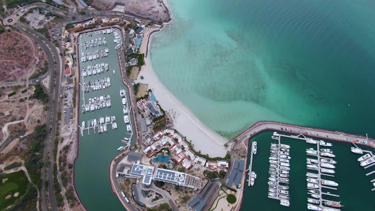 Overhead La Paz frame with the left marina basin centered, rows of docked boats, beach sand stretching toward the lower marina entrance, shallow turquoise water, and no visible people.