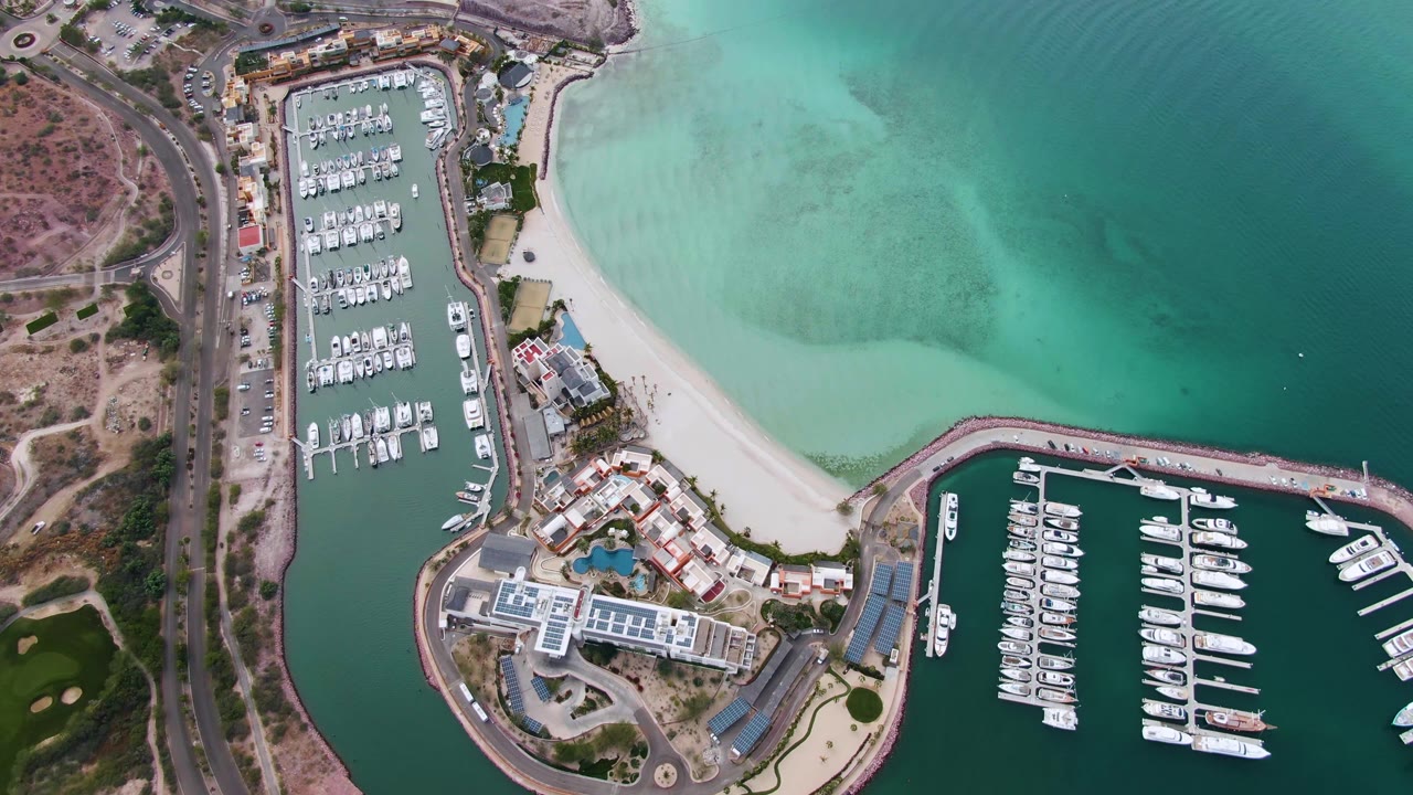 Overhead La Paz frame with the marina basin at left and lower right, a broad sandy shoreline, darker green water offshore, visible vehicles on the road, and no visible people.