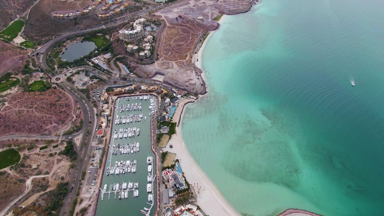 Opening aerial frame over La Paz, Baja California Sur, with a marina full of docked boats beside a sandy beach and turquoise water, no visible people.