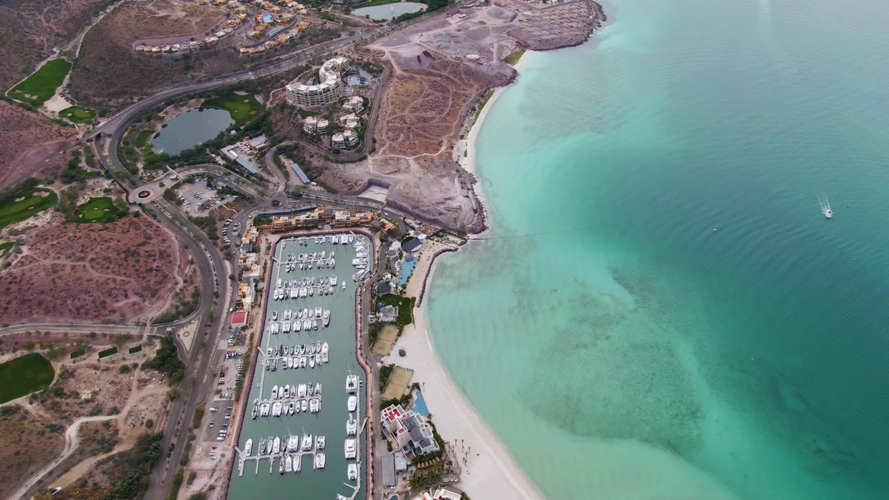 Aerial frame in La Paz showing the marina basin lower left, beach curving along shallow turquoise water, roads inland, and no visible people.