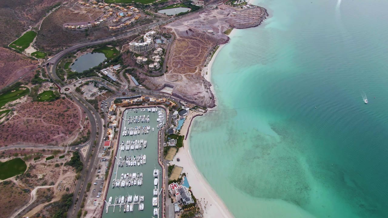 La Paz aerial frame with docked marina boats, a sandy shoreline, pale blue-green water, inland roads and buildings, and a small boat visible offshore, no visible people.