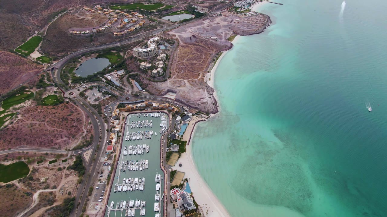 Oblique aerial view of marina, road, beach, and turquoise bay water.