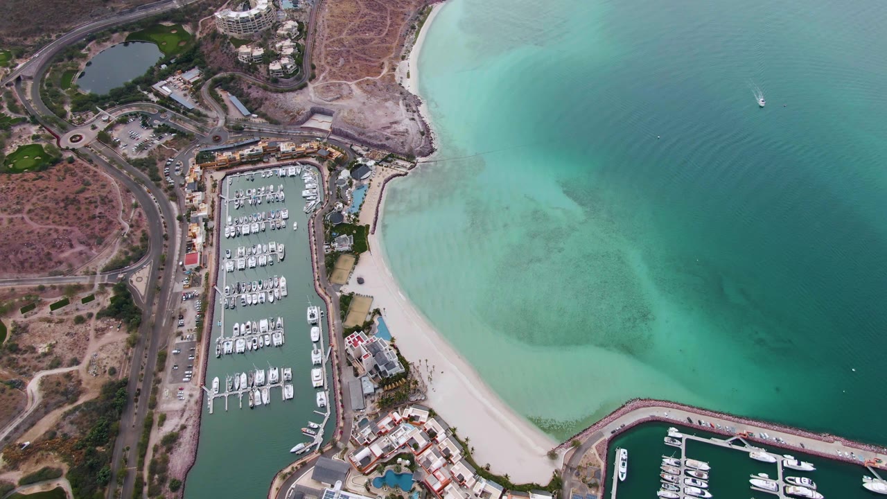 Overhead view of a La Paz marina with rows of docked boats, a pale sandy beach, dry shoreline roads, turquoise coastal water, and no visible people.