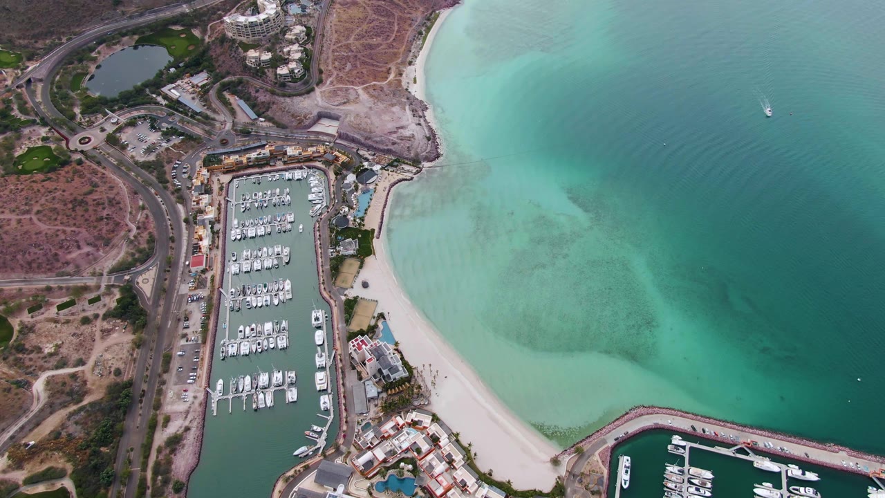 Aerial frame over La Paz with the marina slightly left of center, docked boats in slips, a curved beach, green-blue shallows, and no visible people.