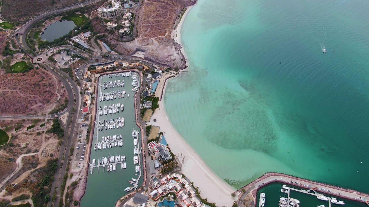High aerial view of La Paz marina, sandy beach, turquoise water, dry coastal terrain, and a small white boat visible offshore with no visible people.