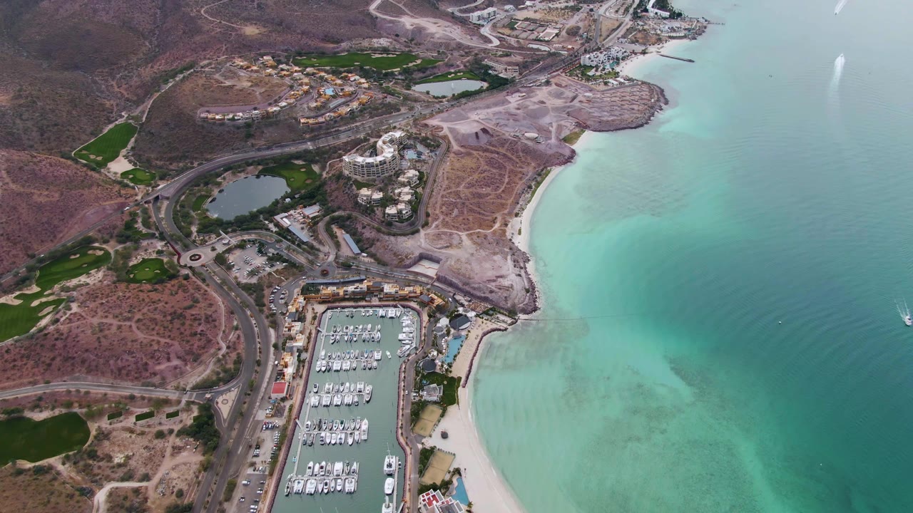 High aerial view over La Paz, Baja California Sur showing a marina with docked boats below, pale beach and turquoise water to the right, dry hills and roads to the left, and no visible people.