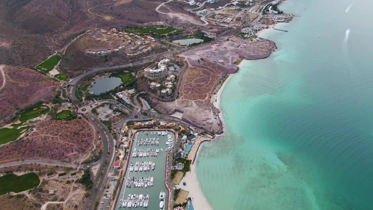 Aerial coastline with marina basin, boats, beach, roads, hills, and shallow water.
