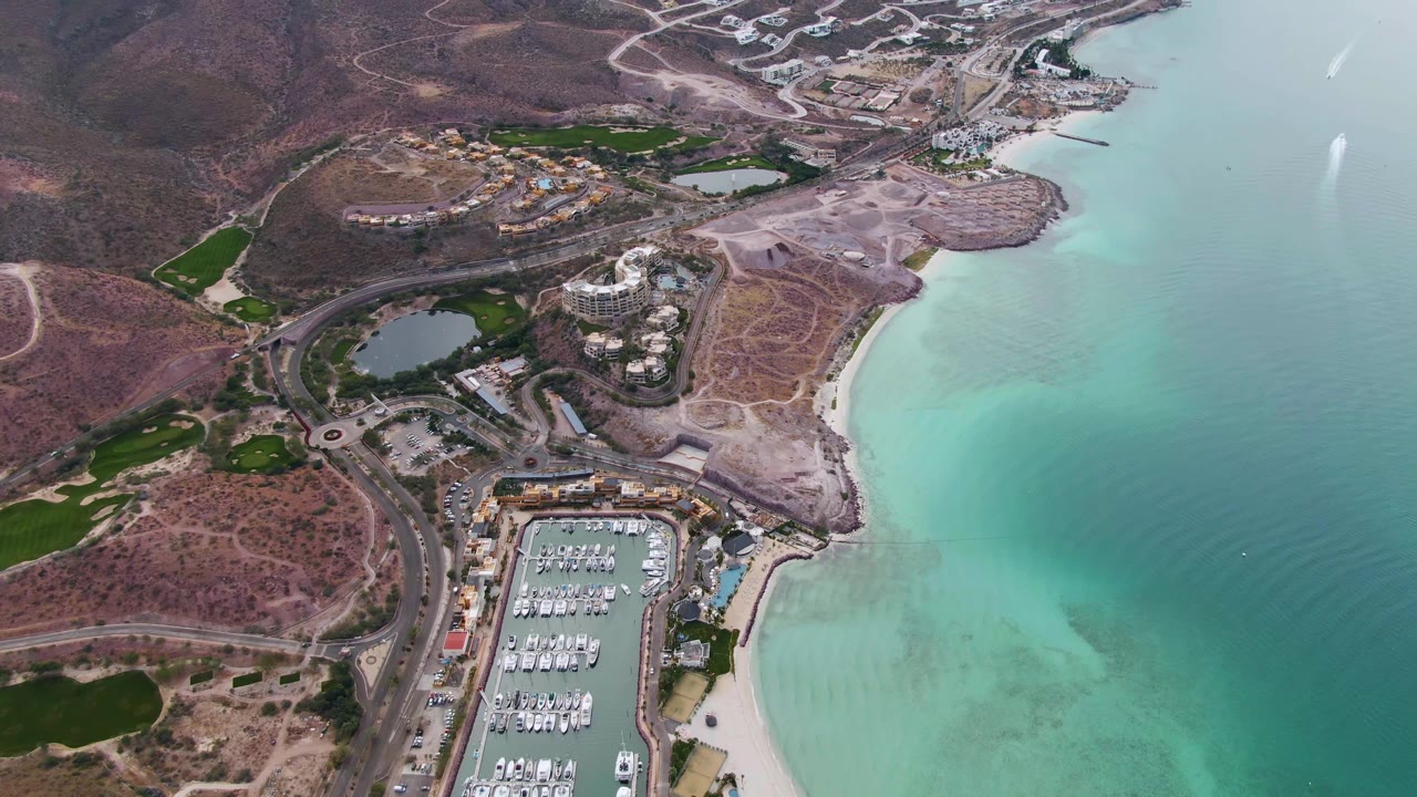 Wide drone view in La Paz, Baja California Sur showing docked boats in the marina at bottom, a sandy shoreline, turquoise bay water with darker patches, dry hills, roads, and no visible people.