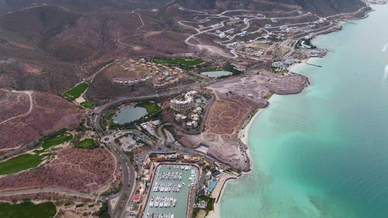 Opening aerial frame over La Paz, Baja California Sur, showing the marina at the lower edge, resort buildings and roads near the beach, turquoise coastal water, dry hills, and no visible people.