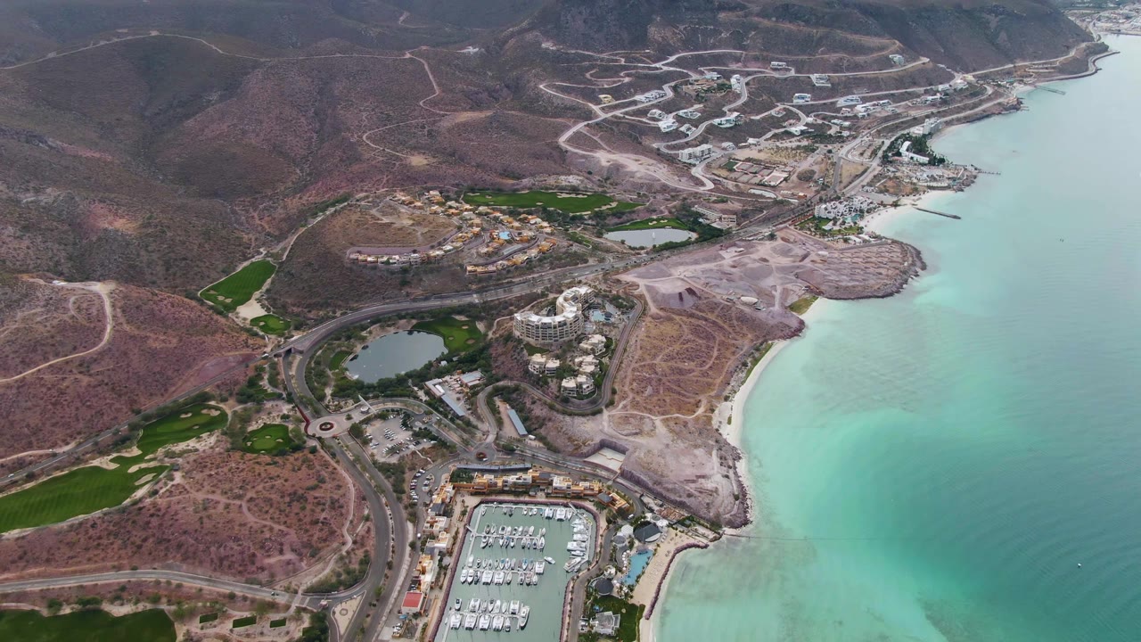 Wide drone view of coastal marina, roads, beach, and mountain-backed resort area.