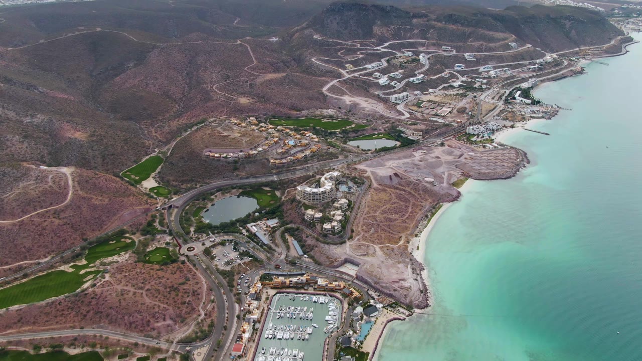 Third aerial frame over La Paz, Baja California Sur, showing the resort and marina area slightly farther along the shoreline, with bright turquoise shallows, road curves, dry hills, and no visible people.