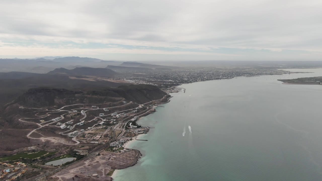 Second frame over La Paz shows the blue-green bay occupying the center, two white water-spray marks on the water, hillside roads at left, distant urban area, and no visible people.