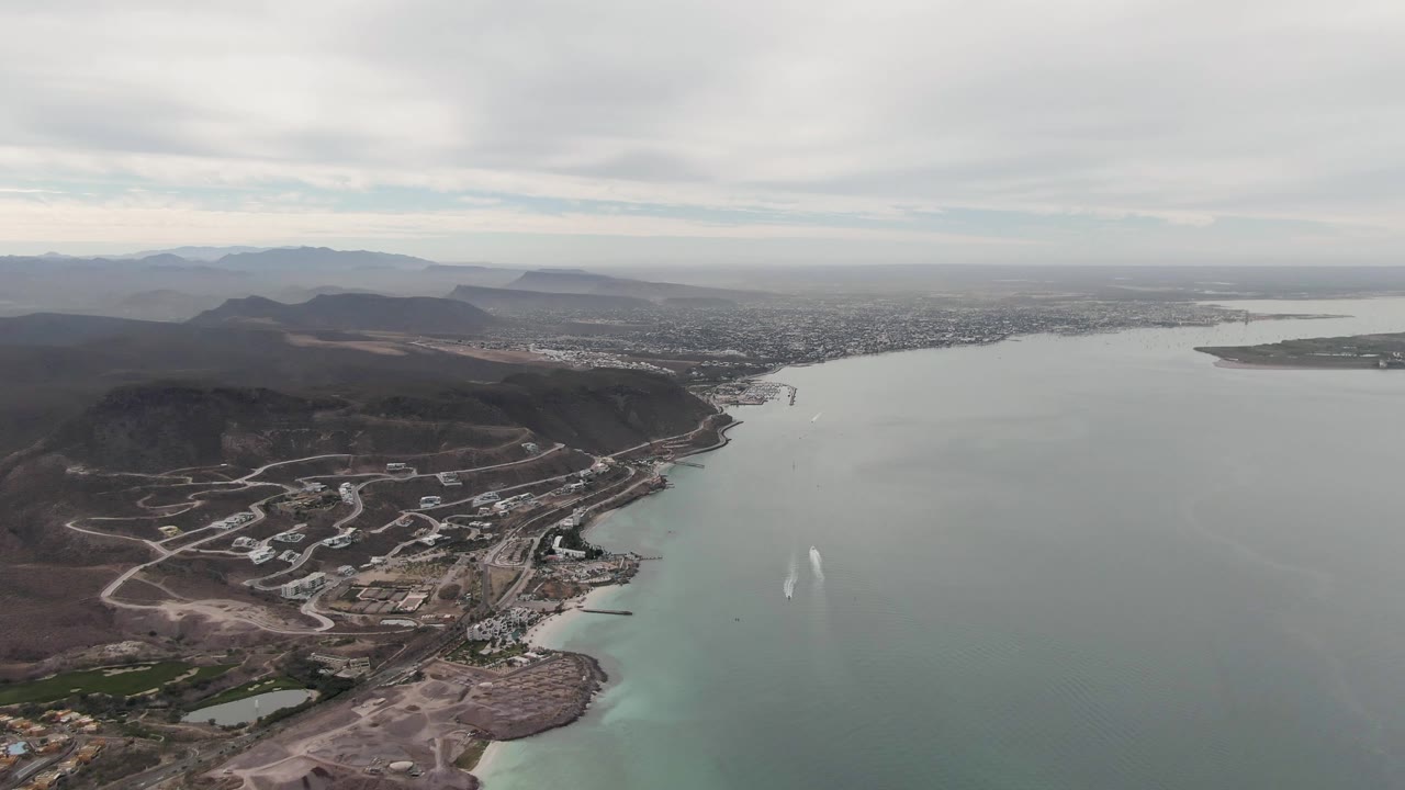 Third frame holds a broad La Paz aerial with the shoreline curving below the hills, pale water extending toward the horizon, distant city development, layered mountains, and no visible people.