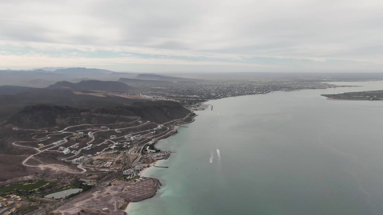 Fourth frame shows a slightly lower-feeling view of La Paz coastline with hillside roads and developed lots more prominent at left, smooth bay water at right, distant city, and no visible people.