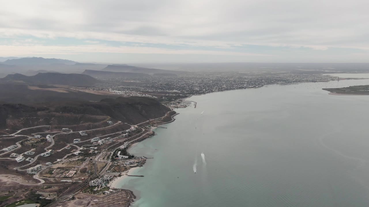Wide aerial view of a coastal waterway, hillside development, city, and distant mountains.