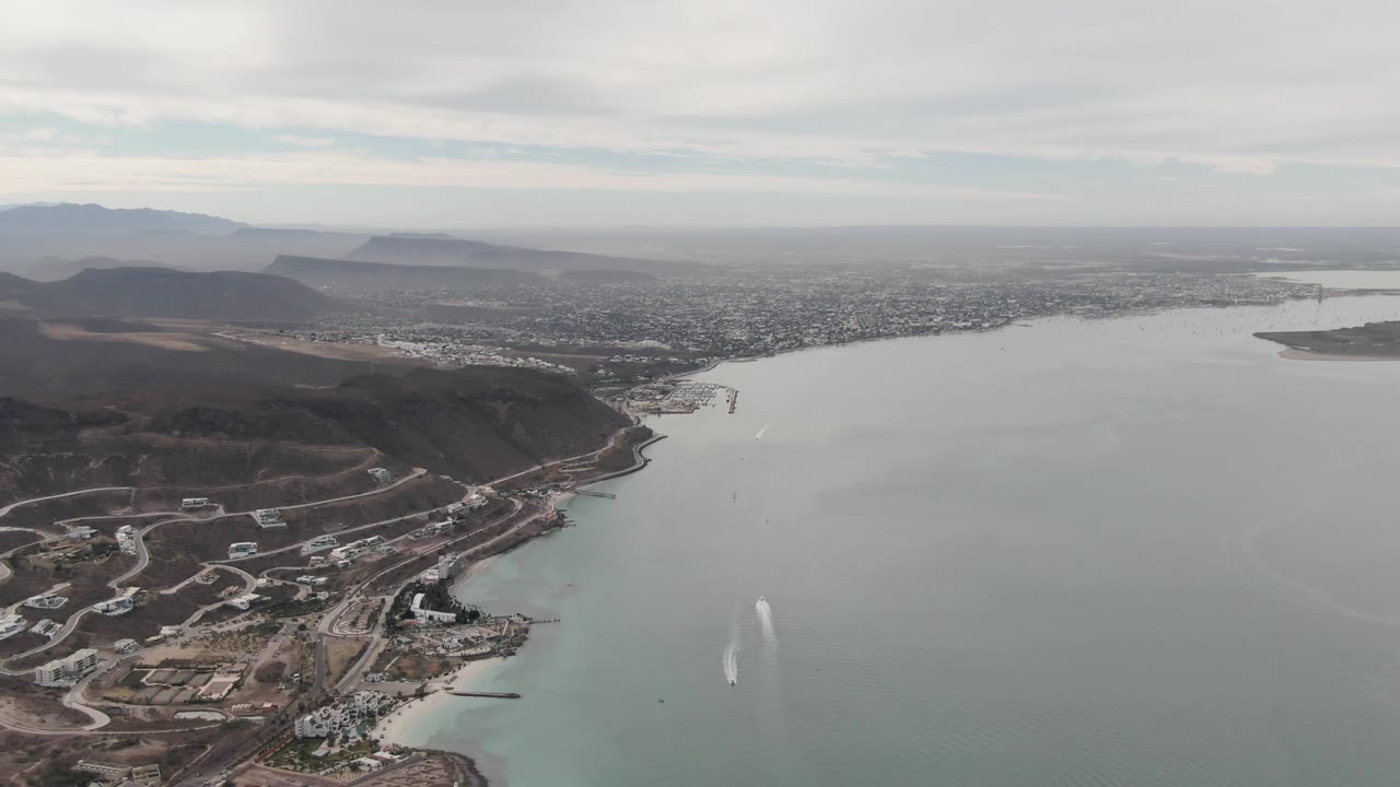 Second aerial frame over La Paz, Baja California Sur, showing the hillside construction area lower in frame, a small white water plume farther out, the city across the water, and no visible people.
