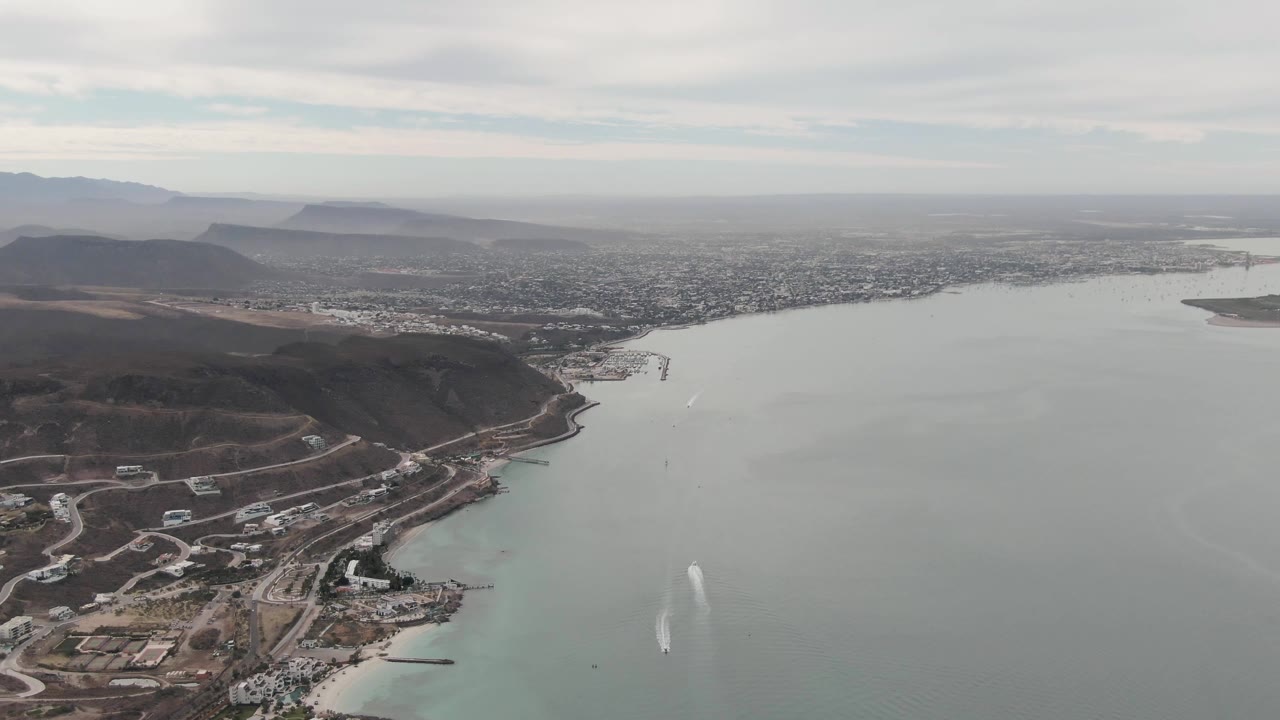 Mid-clip aerial frame of La Paz, Baja California Sur, with the waterway filling most of the image, shoreline construction at the lower edge, city detail across the bay, distant mountains, and no visible people.