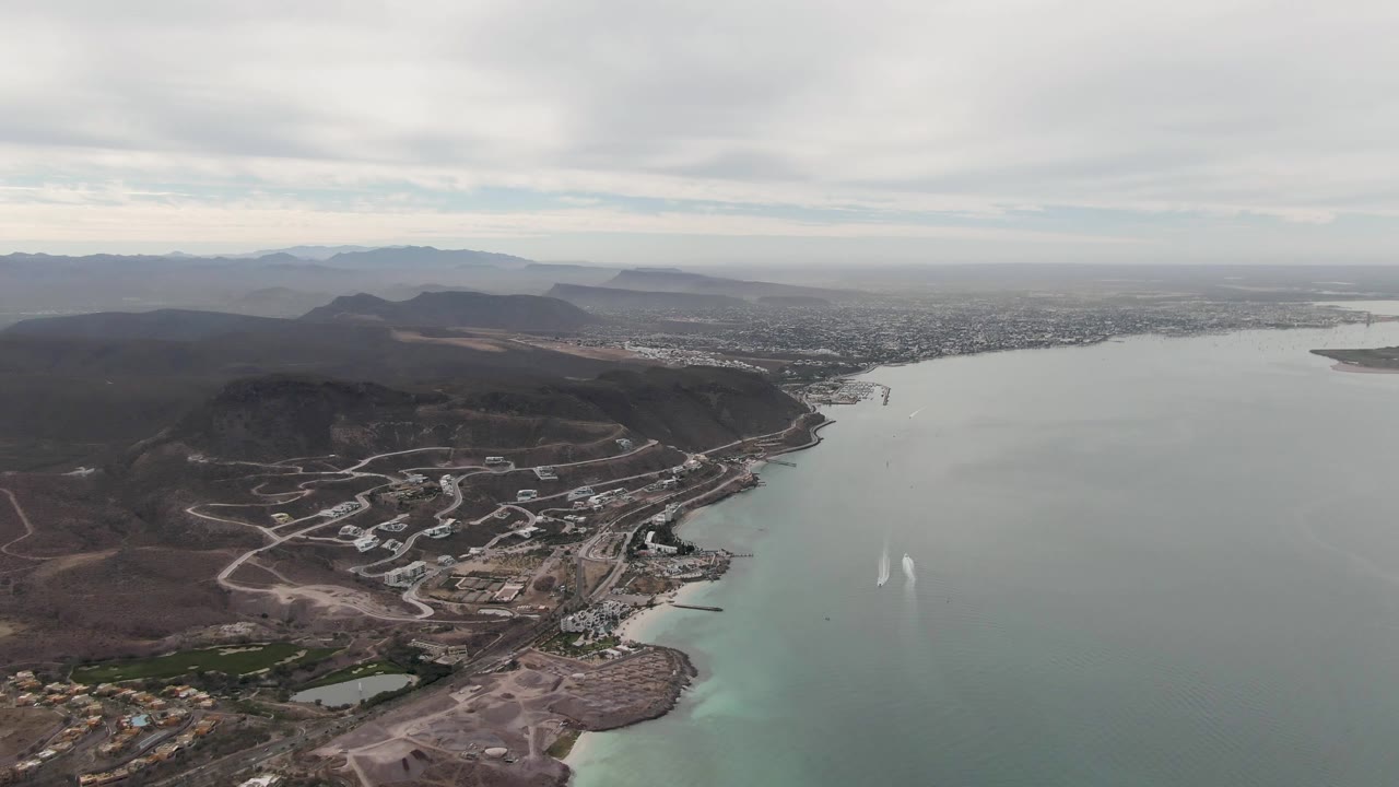 Aerial view over La Paz, Baja California Sur with gray-green bay water, dry hills, shoreline roads, distant city, mountains, overcast sky, and no visible people.