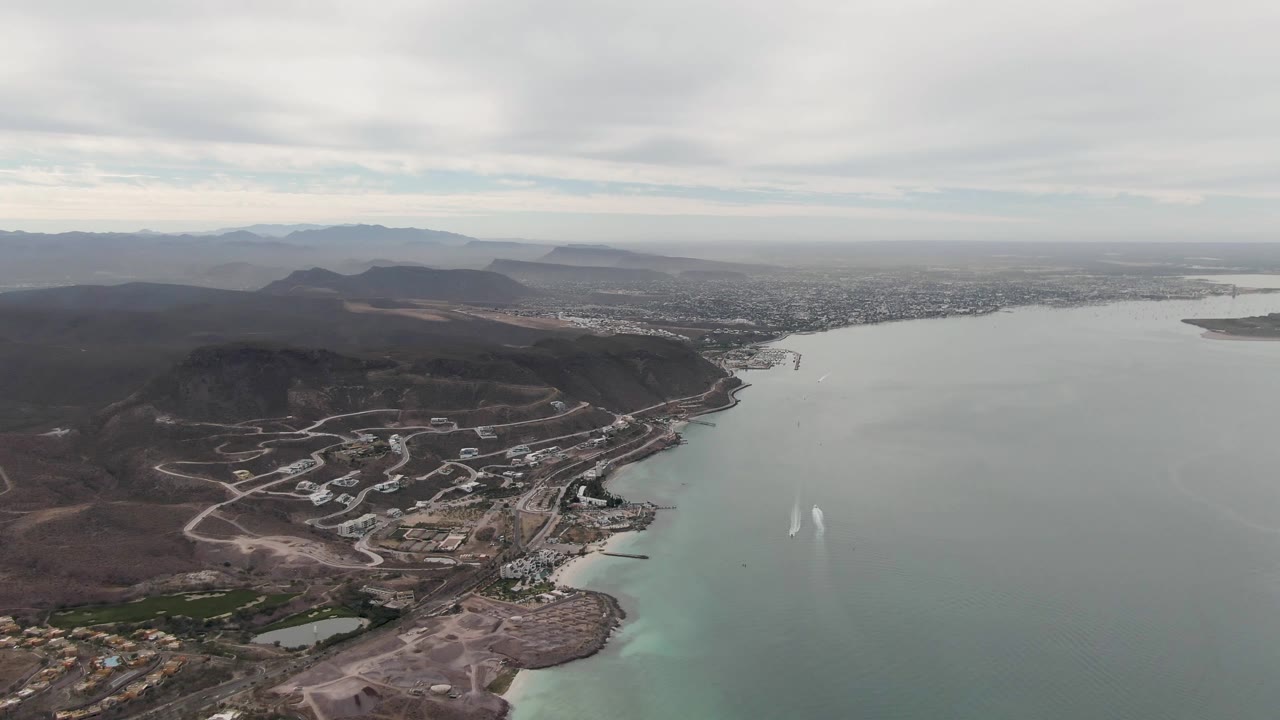Aerial coastal panorama with open water, hillside roads, distant city, and white wake trails.