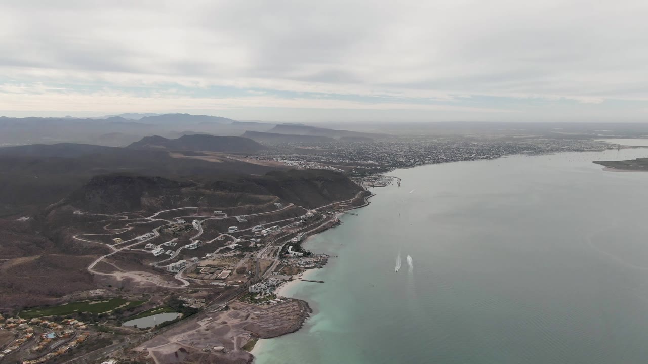 Drone-height view over La Paz with a broad gray bay, twin white wake marks on the water, brown hills with road cuts, distant urban area, mountains, and no visible people.