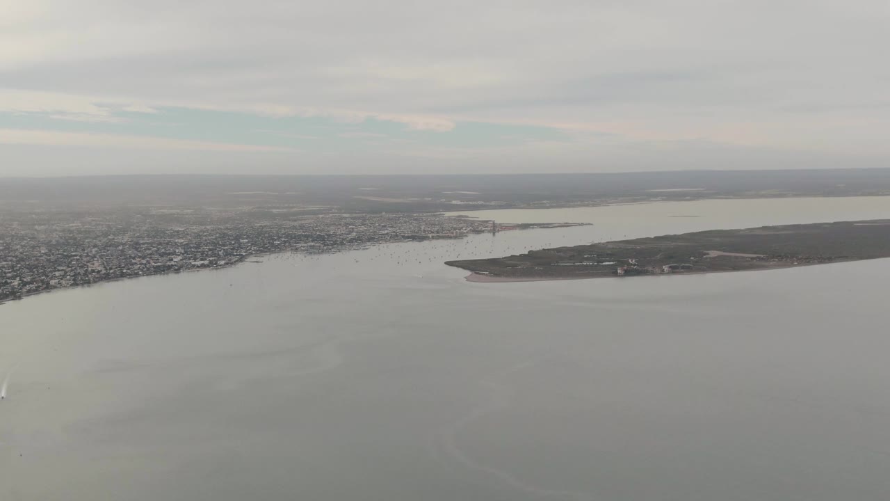 Aerial view over La Paz bay with a faint white boat wake on the water, the island stretching across the middle of frame, city buildings on the far shore, gray clouds, and no visible people.