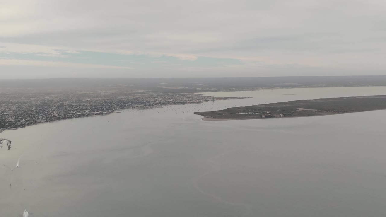 High aerial frame in La Paz, Baja California Sur showing open bay water, a long island, the city shore, a small dock or marina structure near the right side, gray cloud cover, and no visible people.