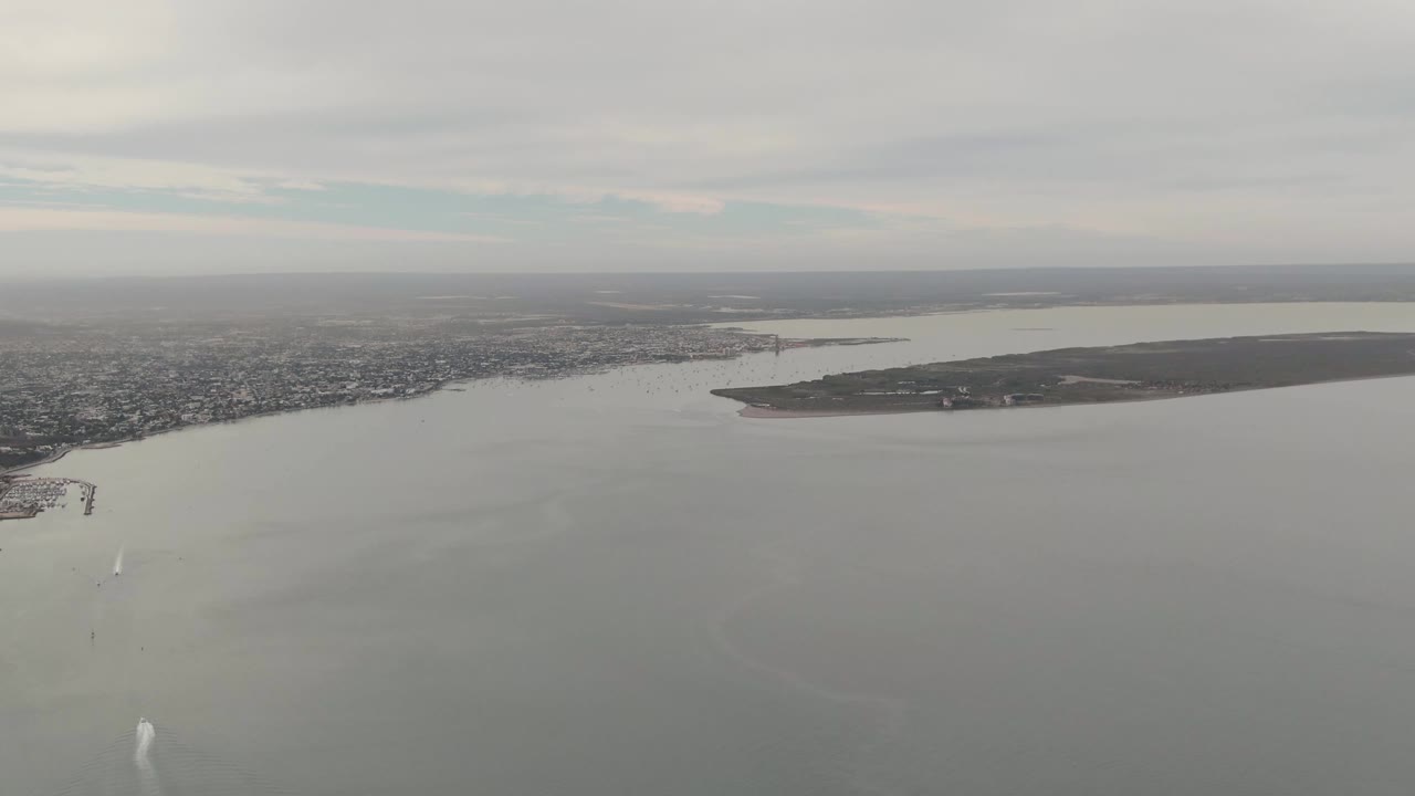 Wide aerial view of La Paz bay with the island centered across the water, city shoreline in the distance, a small white wake near the lower left, soft gray light, and no visible people.