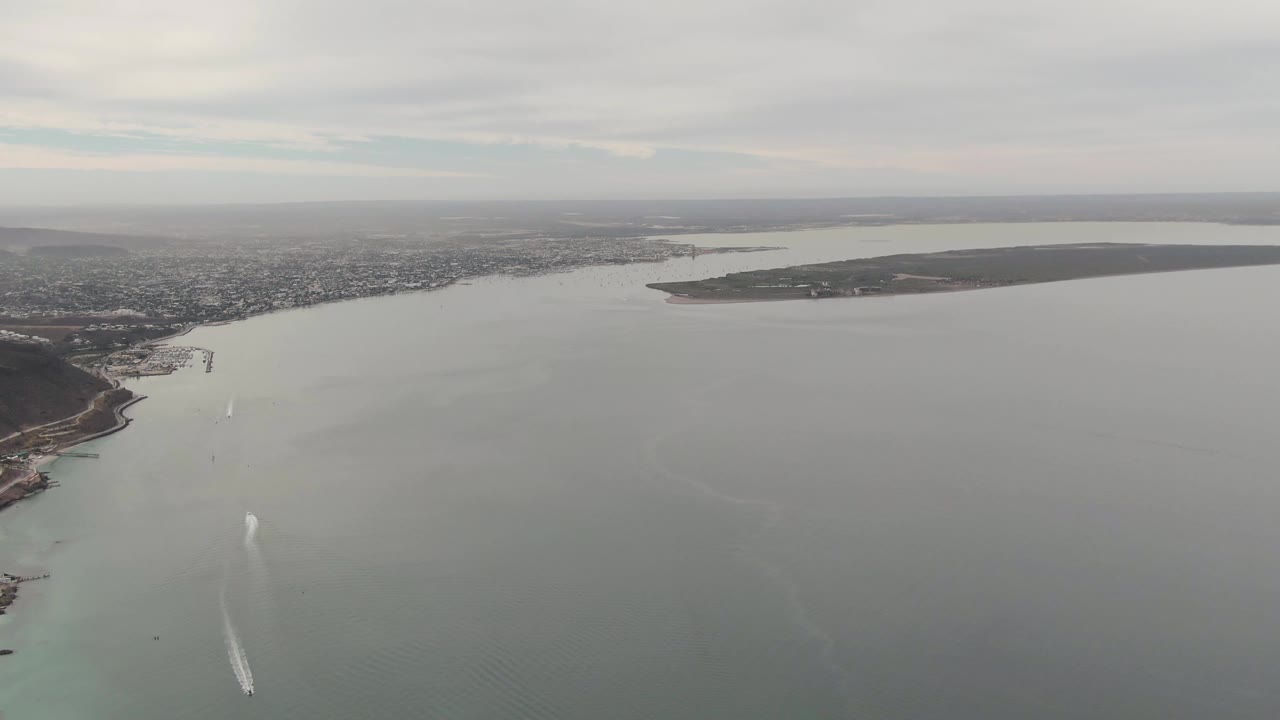 Aerial view over La Paz, Baja California Sur, with gray-green water, distant city shoreline, a long landmass on the horizon, and a small boat wake near the lower left, with no visible people.