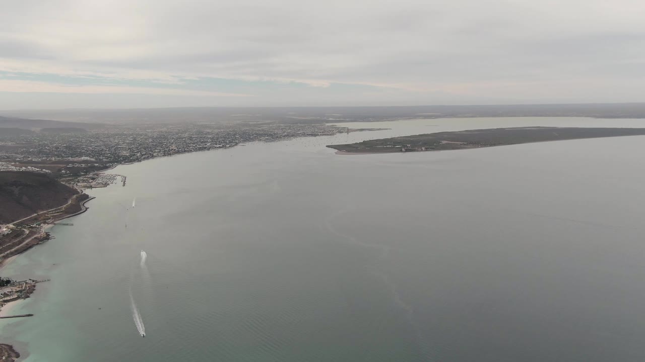 High aerial frame over La Paz showing open water in the foreground, a dark hilly shoreline at right, distant low city development, and a white boat wake moving through the bay, with no visible people.